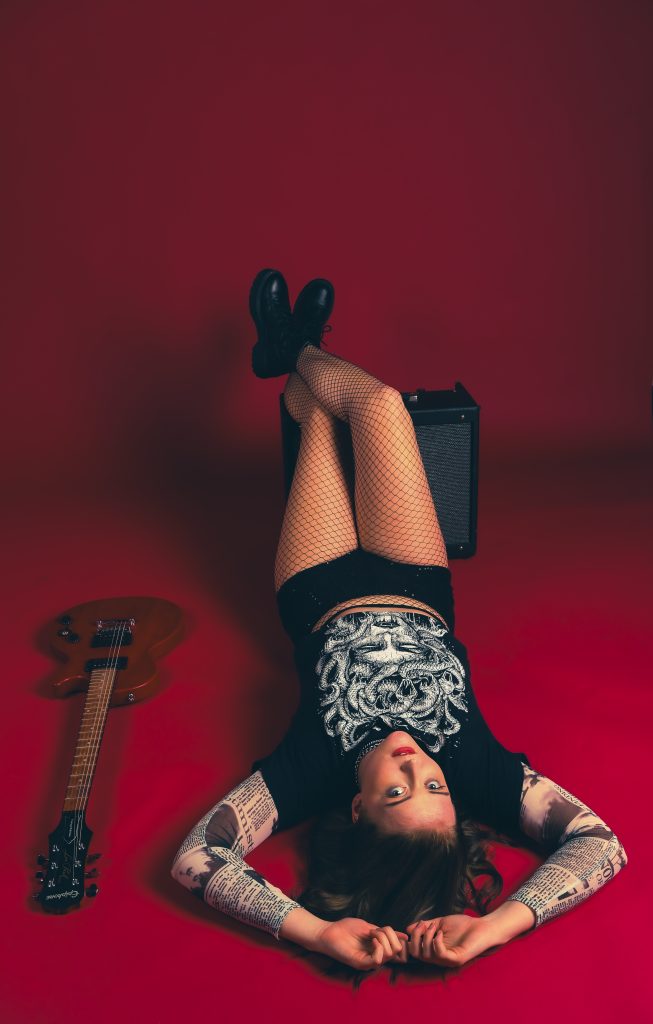 Musician Alicia poses laying on a red seamless backdrop paper with her guitar to her left.